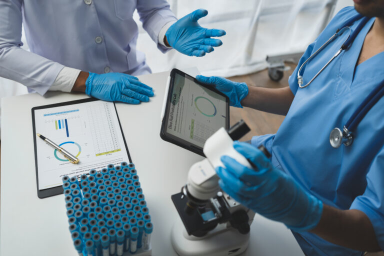 Two healthcare workers in blue gloves discussing medical data displayed on a tablet in a clinical laboratory.